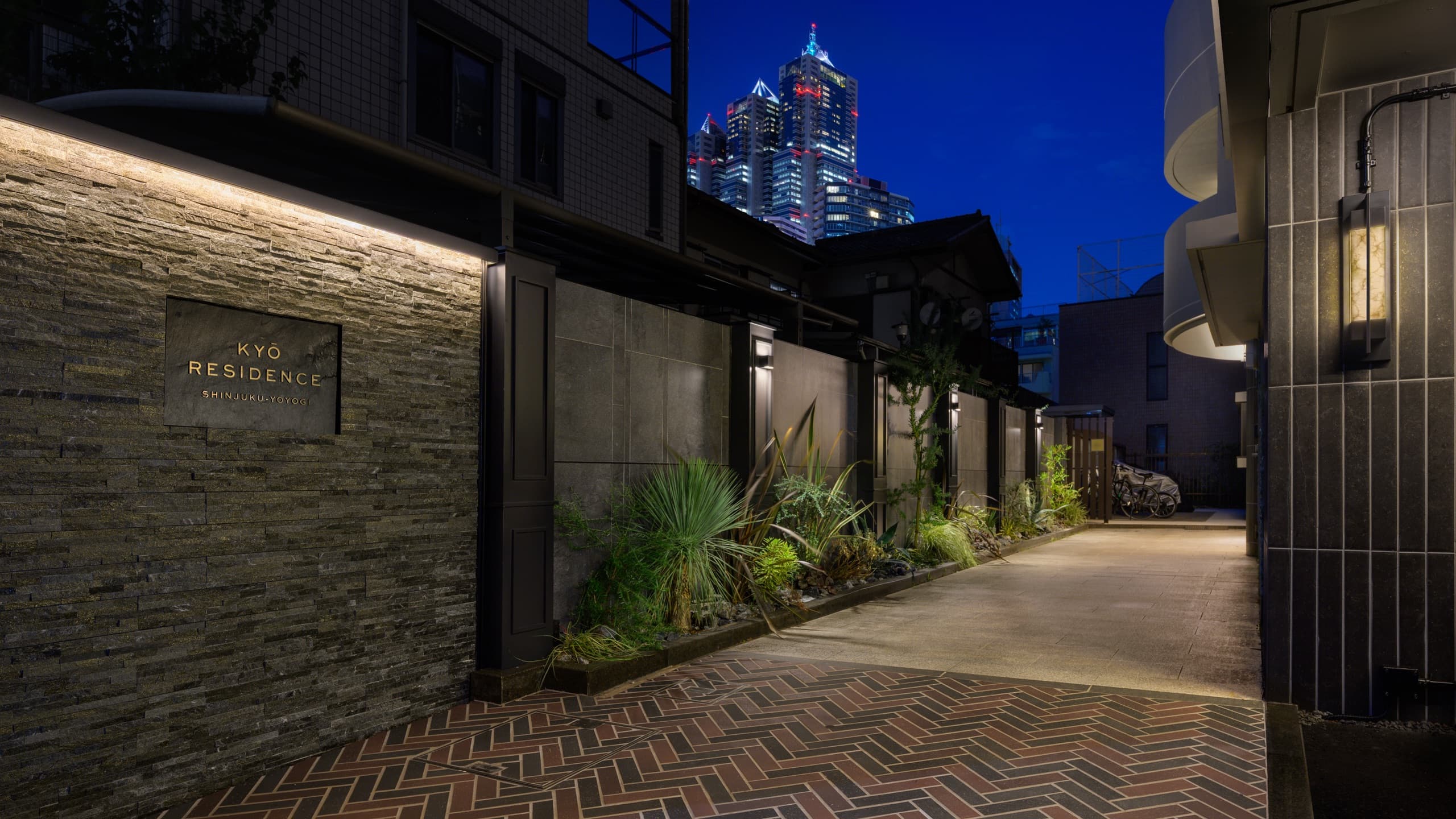 KYO Residence エントランス — 夜のアプローチ、石壁のサインと新宿の高層ビル